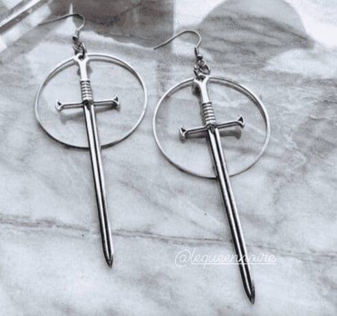 A pair of silver earrings shaped like swords, each with a long, pointed blade and a cross-guard hilt. The swords are suspended within a circular hoop, creating a striking and edgy design. The earrings are laid out on a marble surface, showcasing their metallic sheen. The image also includes the handle '@lequeennoire' in white script, positioned near the bottom right corner.